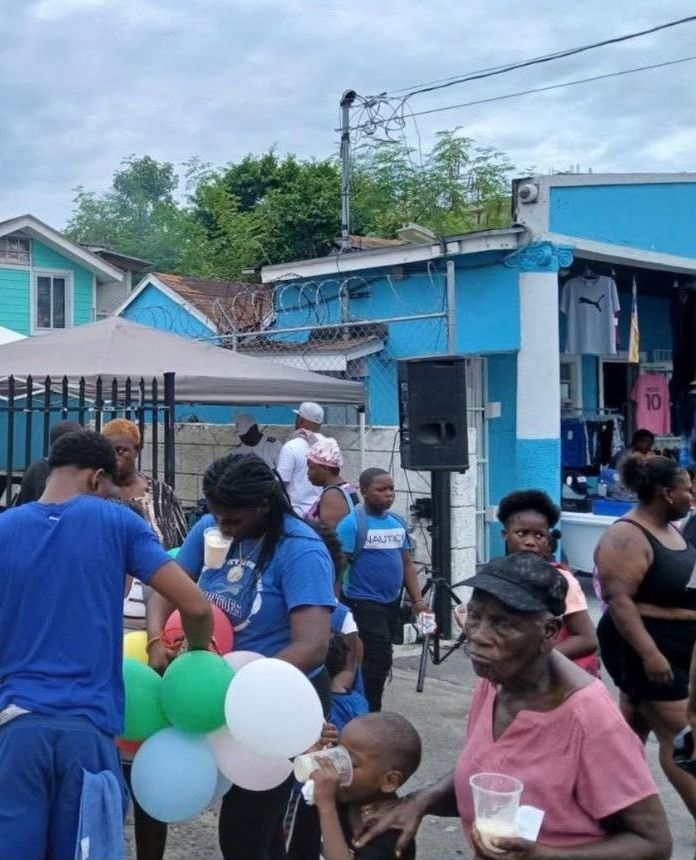 Children and adults gather in the street holding cups and balloons near a blue building at the event