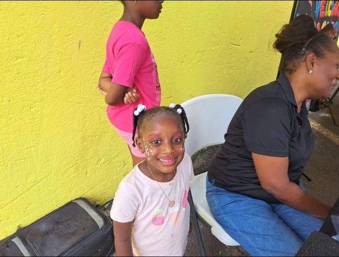 A smiling girl with face paint sits in front of a yellow wall at the Back to School event