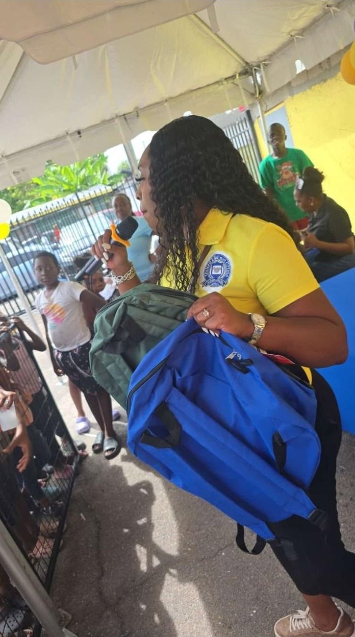 A woman in a yellow UAC polo holds up a blue backpack as children gather at the giveaway