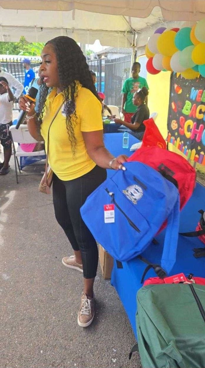 A woman in a yellow polo holds a microphone and blue and red backpacks under a Back to School tent