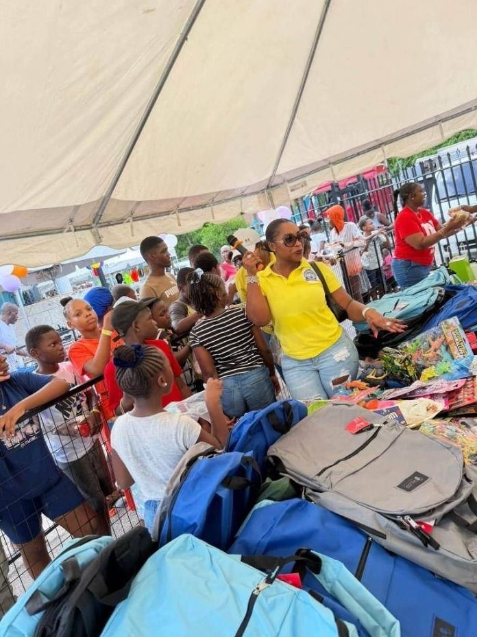 A woman in a yellow UAC polo stands among children beside a table piled with new backpacks