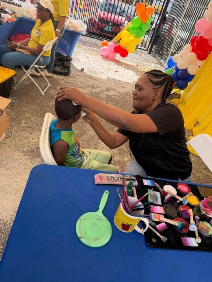 A woman paints a young child's face at a table of face paints and brushes under a tent