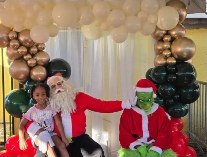 A smiling girl sits beside Santa and a costumed Grinch under a balloon arch at the Christmas event