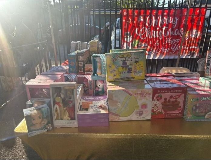 Boxed dolls, craft kits and games displayed on a gold-covered table beneath a Merry Christmas banner
