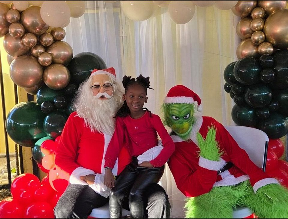A smiling girl in red sits on Santa's lap beside a costumed Grinch under a balloon arch