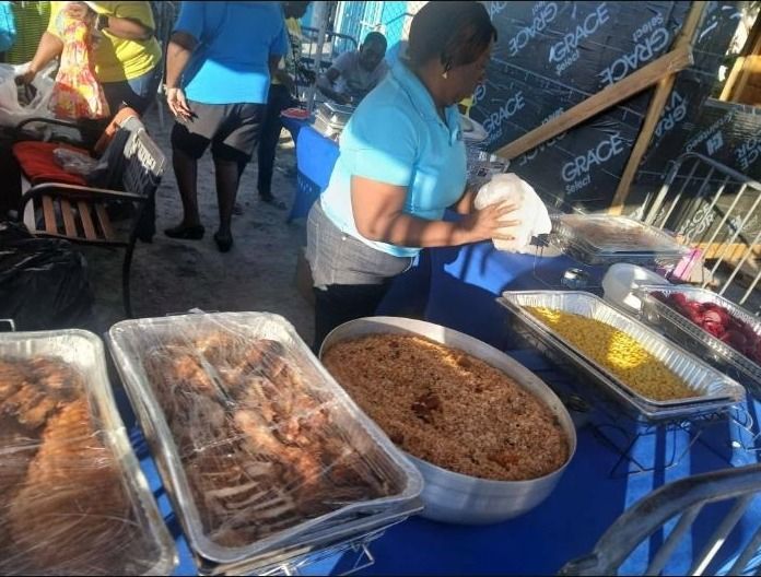 A woman in a light blue shirt serves from trays of baked chicken, rice and corn on a blue table