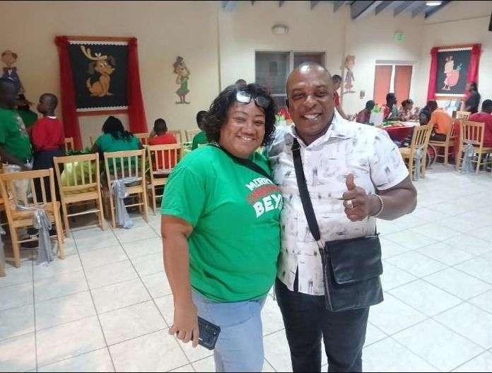 A woman in a green Christmas shirt and a man pose together in a hall decorated for a Christmas party