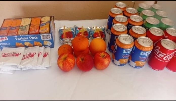 Crackers, apples, oranges, juice boxes and canned drinks arranged on a white table as a snack donation
