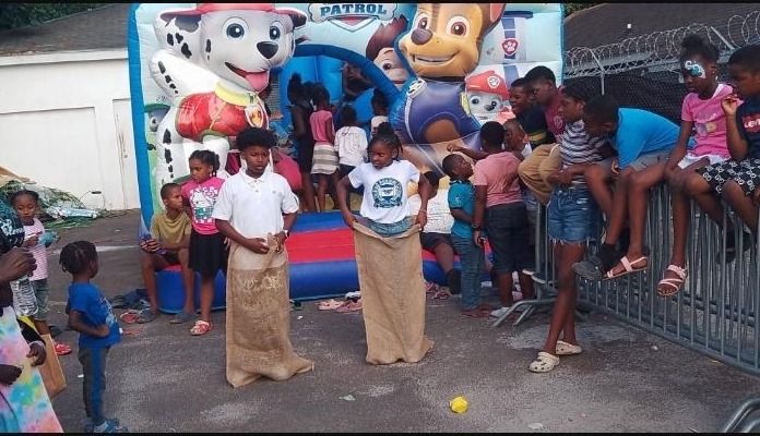 Two children race in burlap sacks while other children watch beside a Paw Patrol bounce house