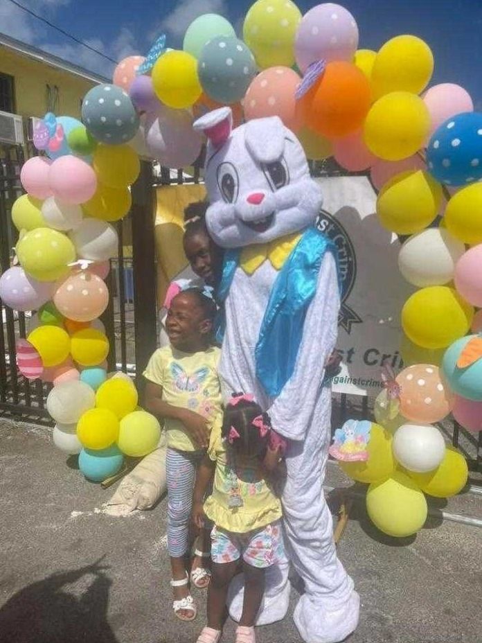 Two young girls and a costumed Easter Bunny pose in front of a pastel balloon arch at the gate