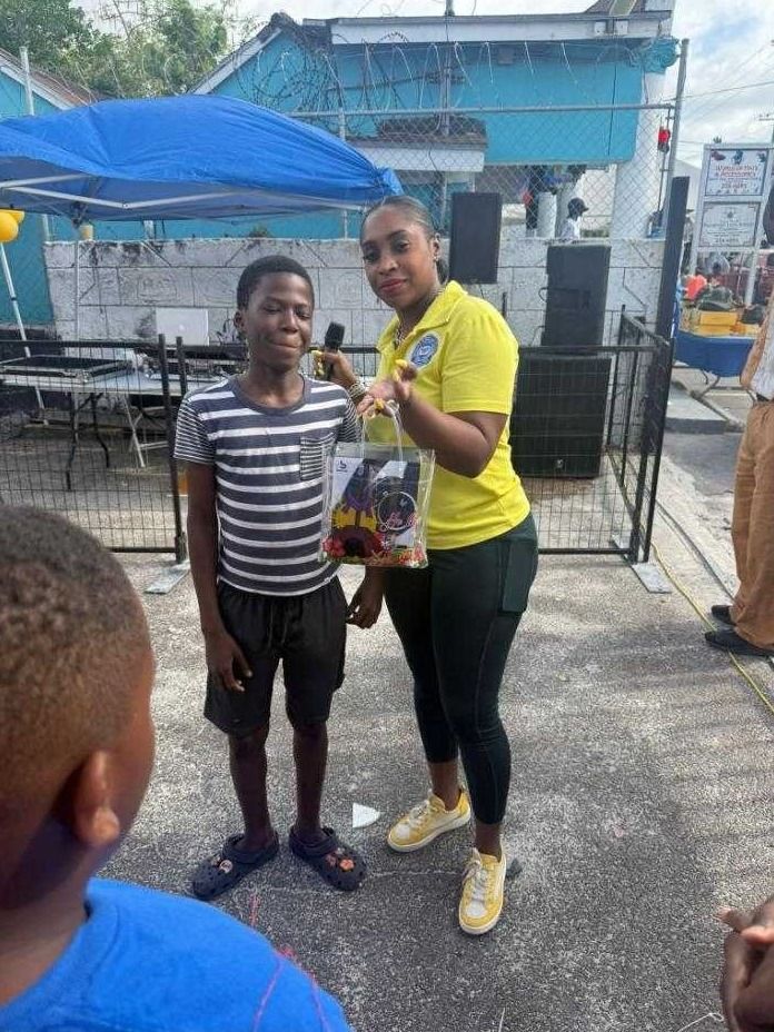 A woman in a yellow polo with a microphone holds a clear gift bag beside a boy