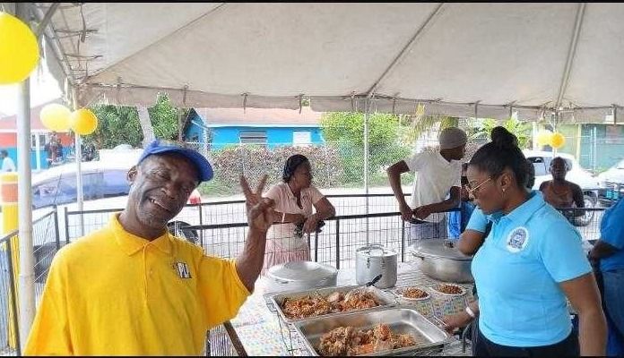 A smiling man in a yellow shirt makes a peace sign beside trays of hot food and servers