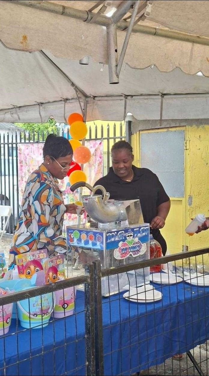 Two women prepare shaved ice cones at an ice cone machine beside Easter buckets