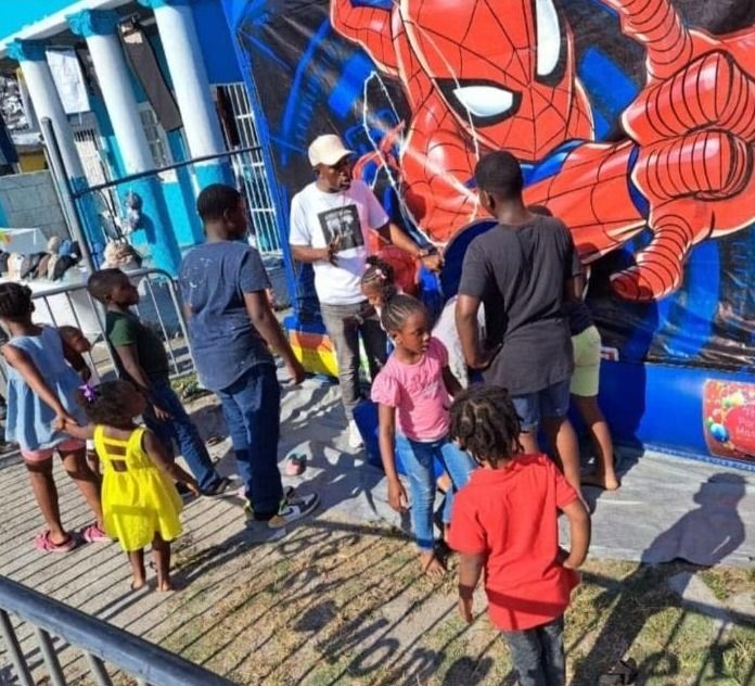Children gather around a man in a white cap in front of a Spider-Man bounce house