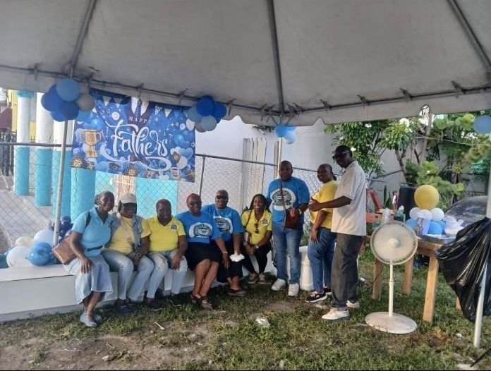 Volunteers in yellow and blue shirts sit and stand under a tent with a Happy Father's Day banner