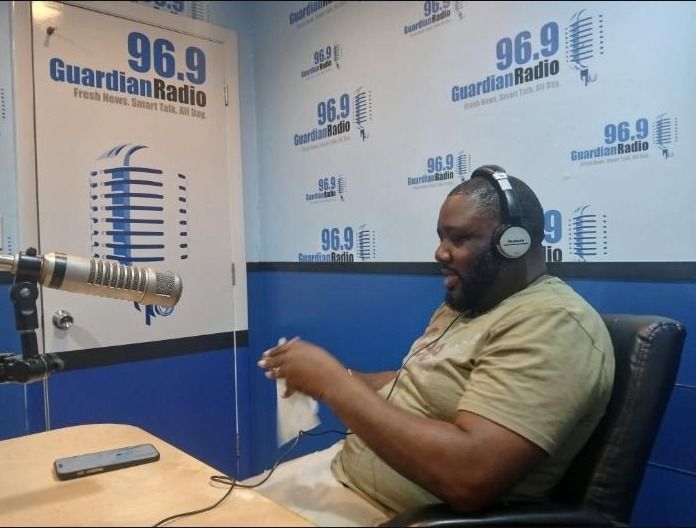 A bearded man in headphones sits at a microphone in the Guardian Radio 96.9 studio