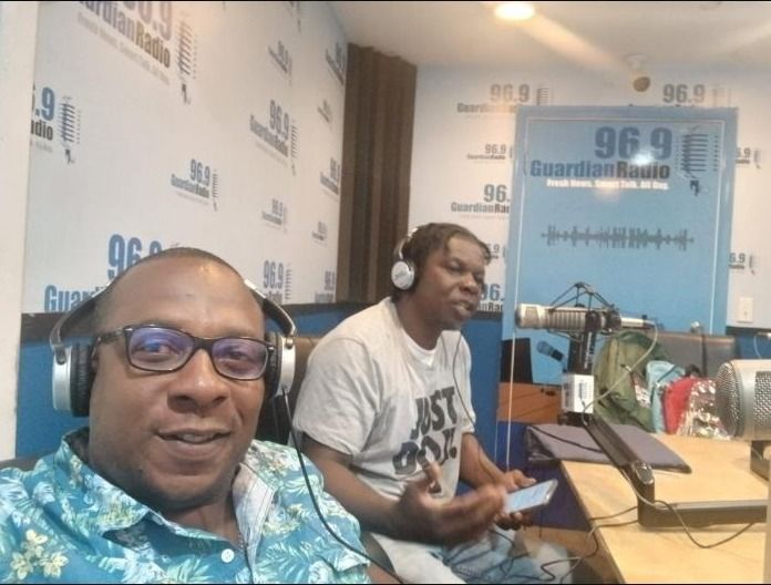 Two men in headphones sit at microphones in front of Guardian Radio 96.9 branded walls