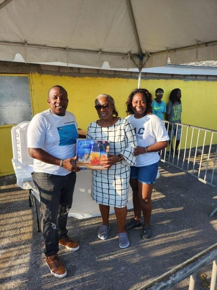 Two volunteers in white shirts stand with a woman in a checked dress holding a boxed glassware set