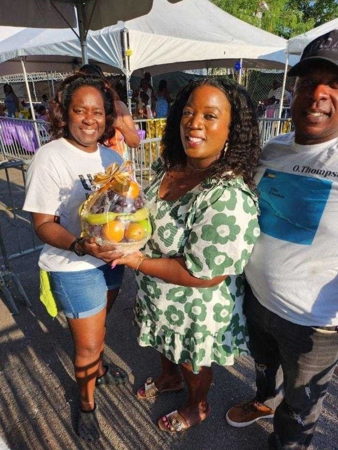 Two volunteers and a woman in a green floral dress hold a fruit gift basket at a barrier
