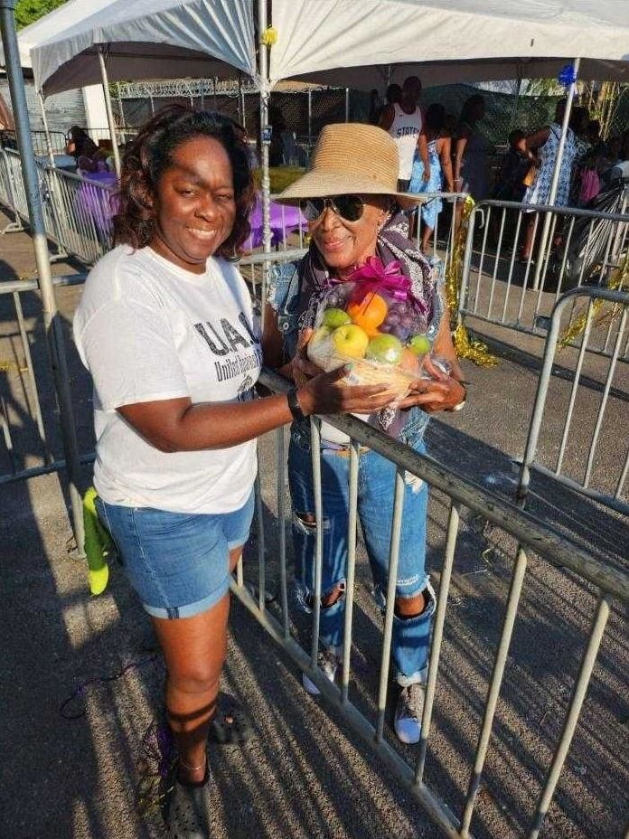 A volunteer and a woman in a straw hat hold a fruit gift basket at a metal barrier