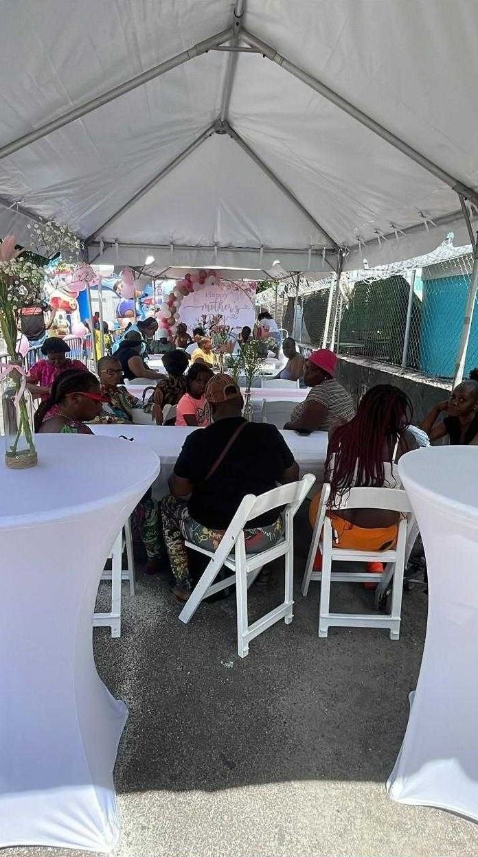 Guests sit at white tables with flower vases under a tent with a Happy Mother's Day balloon display