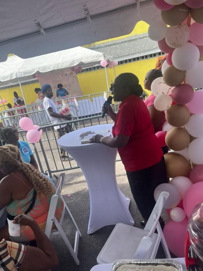 A woman in a red shirt speaks into a microphone beside a table and a balloon garland
