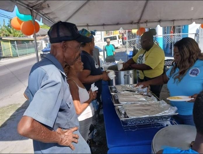 An older man in a gray shirt waits in line as volunteers serve food from foil trays