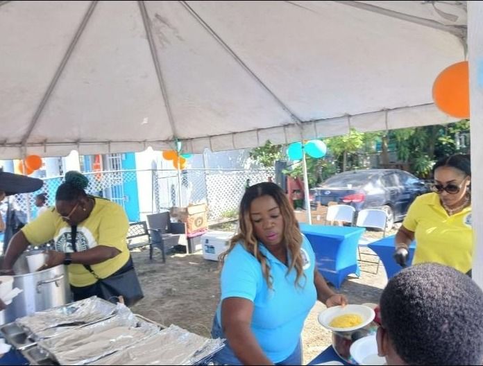 Two women in light blue and yellow shirts serve plates of scrambled eggs from a serving table under a tent