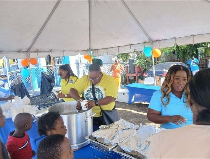 Volunteers in yellow and light blue shirts serve hot food from a large pot and foil trays to children