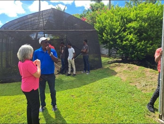 A guide in a blue shirt and a woman in pink talk while young visitors stand near a shade house